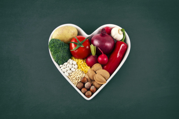 A white heart-shaped bowl full of brightly colored, healthy vegetables, nuts, beans, and more. 