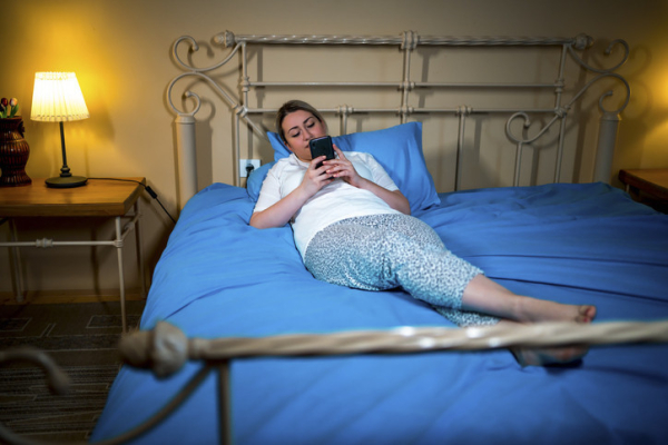 A woman in nightwear looking at her smartphone white lying on top of a bed with blue sheets and pillowcases, bedside lamp on